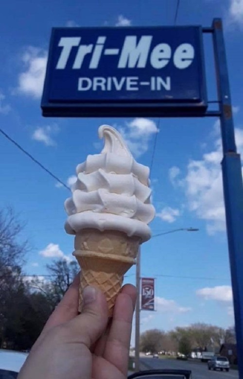 A hand holds a soft-serve ice cream cone in front of a blue sign that reads "Tri-Mee Drive-In" against a cloudy sky.