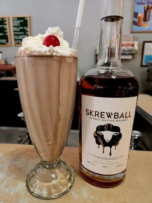 A tall glass of chocolate milkshake topped with whipped cream and a cherry, next to a bottle of Skrewball peanut butter whiskey.