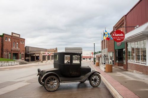 A vintage car parked on a quiet street lined with brick buildings and flags, under a cloudy sky.