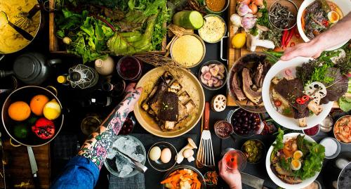 A vibrant spread of various dishes, fresh ingredients, and drinks, with hands serving food and colorful garnishes.