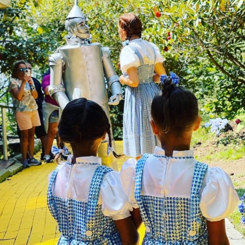 Two girls in blue dresses watch as Dorothy interacts with the Tin Man on a yellow brick road, surrounded by greenery.