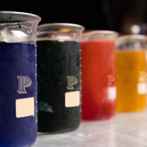 Four beakers filled with colorful liquids: blue, green, red, and yellow, lined up on a lab table.