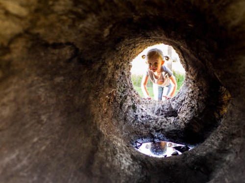 A child peeks through a circular hole in a structure, with sunlight illuminating their face and a reflection in water below.