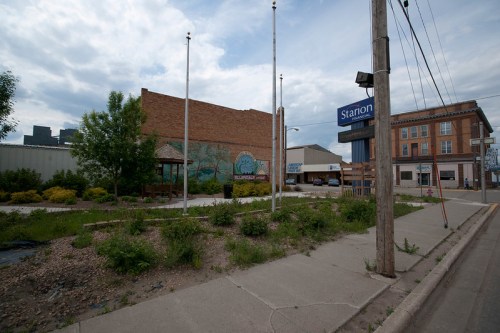A street view featuring a mural on a building, flag poles, and a sign for "Starion" amidst greenery and cloudy skies.