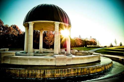 A gazebo with a dome and columns, surrounded by a fountain, illuminated by sunlight in a serene park setting.