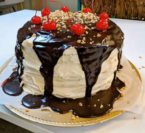 A tall cake with white frosting, topped with chocolate drizzle, chopped nuts, and maraschino cherries.
