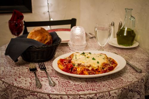 A plate of lasagna topped with cheese, served with a bread basket, water, and olive oil on a lace-covered table.