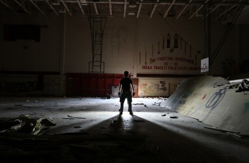 A silhouette of a person stands in a dark, abandoned gym, surrounded by debris and graffiti on the walls.