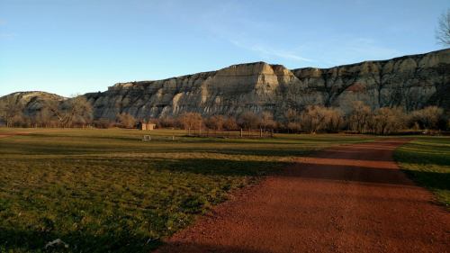 A scenic view of a grassy area with a dirt path leading to a large, layered rock formation under a clear blue sky.