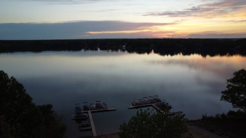 A serene lake at sunset, with boats docked and colorful reflections on the water. Trees frame the scene.