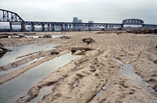 A sandy riverbank with exposed mud and water pools, featuring a large bridge and city skyline in the background.