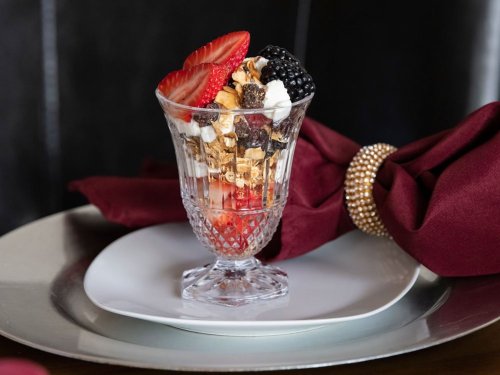 A glass dessert cup filled with layers of granola, berries, and cream, placed on a white plate with a burgundy napkin.
