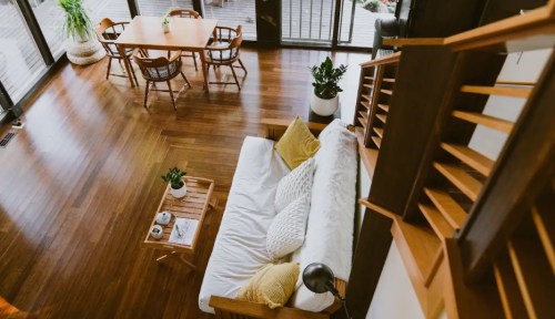 A cozy living room with a white sofa, wooden furniture, and a dining table, featuring large windows and plants.