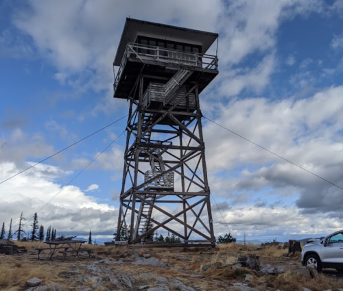 A tall wooden fire lookout tower stands against a cloudy sky, surrounded by grassy terrain and a parked vehicle nearby.