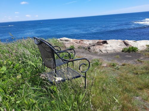 A black metal bench sits on grassy rocks overlooking a calm blue ocean under a clear sky.