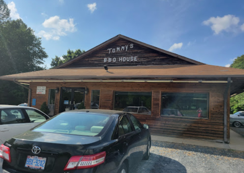 A wooden building with a sign reading "Tommy's B-B-Q House," surrounded by parked cars and trees under a blue sky.