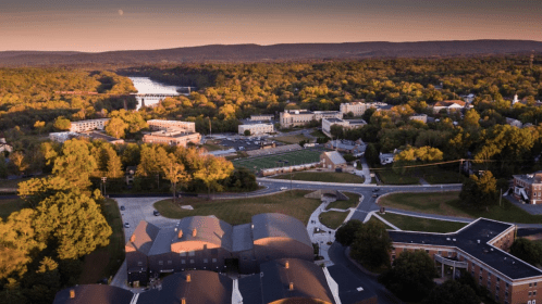 Aerial view of a scenic landscape with buildings, trees, and a river at sunset, surrounded by rolling hills.