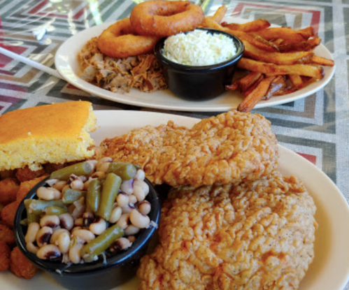 A plate of fried chicken, black-eyed peas, cornbread, and onion rings, with a side of fries and coleslaw in the background.