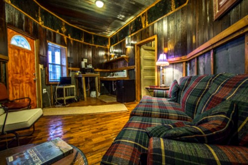Cozy room with wooden walls, a plaid sofa, a lamp, and a small kitchenette, featuring warm lighting and rustic decor.