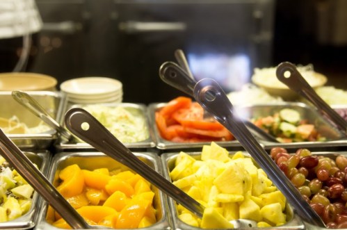 A variety of fresh fruits and salads displayed in metal trays with serving tongs.