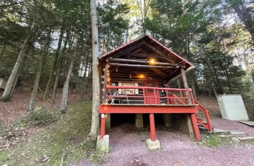 A cozy wooden cabin with a red porch, surrounded by trees and a gravel path. Warm lights adorn the exterior.