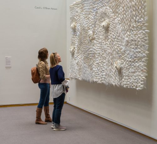 Two women admire a large, textured white artwork on a gallery wall.