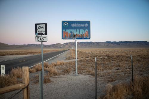 A welcome sign for Nevada beside a deserted road, with mountains in the background and a clear sky at dusk.