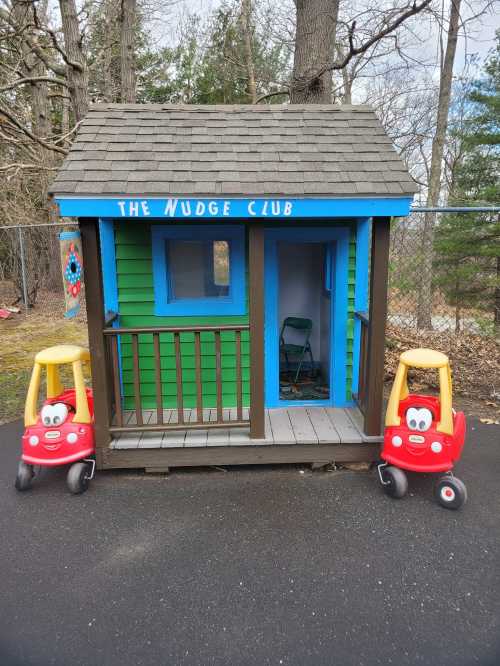 A colorful playhouse named "The Nudge Club" with two red toy cars outside, set in a wooded area.