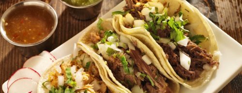 A plate of three tacos filled with meat, topped with onions and cilantro, served with salsa and radishes on the side.