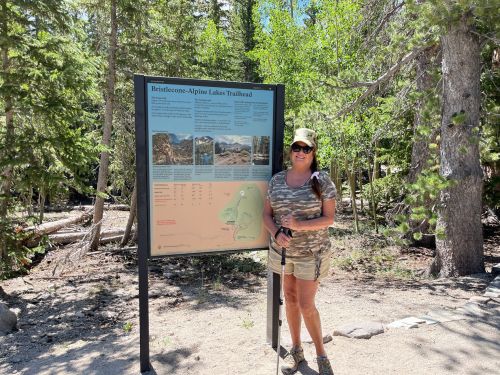 A woman stands next to a trailhead sign in a forested area, holding a hiking pole and wearing a hat and camouflage outfit.
