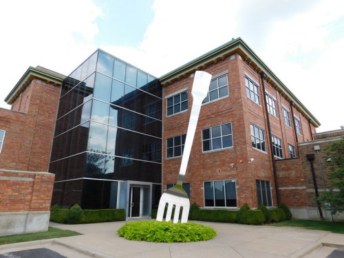A large fork sculpture stands in front of a modern brick building with glass windows and green landscaping.