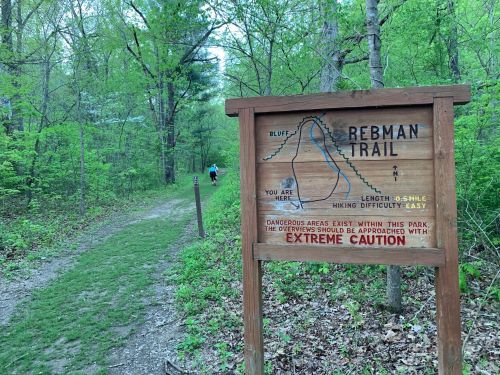 A wooden sign for Redman Trail, indicating location and difficulty, with a hiker on the path surrounded by greenery.