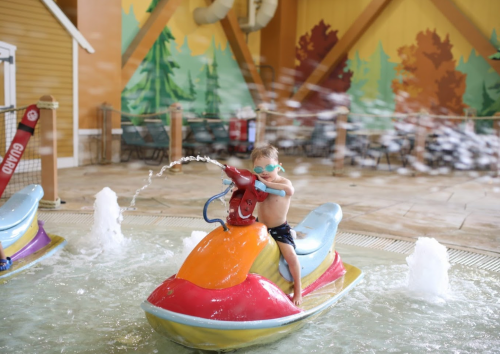 A child in swim goggles plays on a colorful water ride, splashing in a pool with a fun, vibrant background.