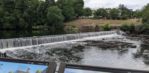 A serene view of a waterfall cascading over rocks, surrounded by greenery and informational signs in the foreground.