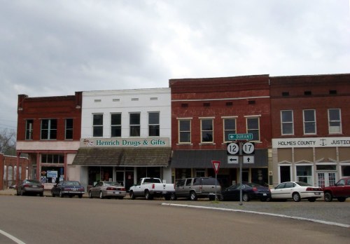 A small town street featuring brick storefronts, including a drugstore, with parked cars and cloudy skies above.