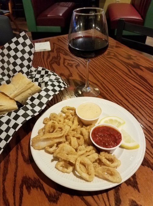 A plate of fried calamari with dipping sauces, lemon slices, a glass of red wine, and a basket of bread on a wooden table.
