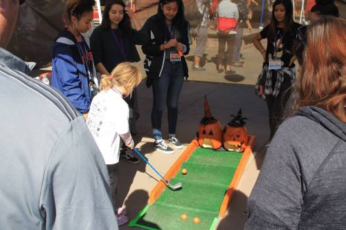 A child plays mini-golf while a group of onlookers watches, with Halloween decorations in the background.