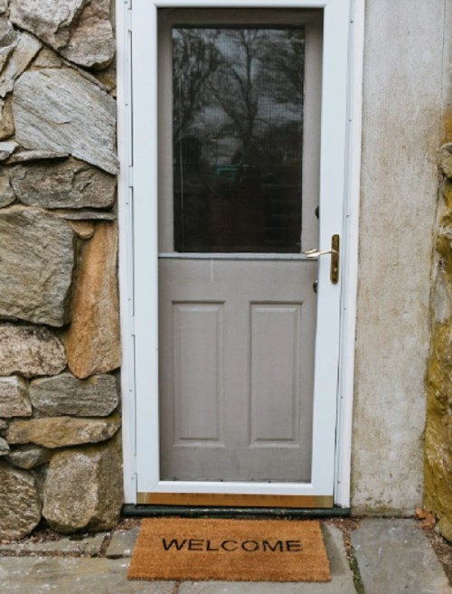 A gray door with a glass panel, framed by stone walls, and a "WELCOME" doormat at the entrance.