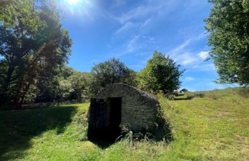 A small stone structure sits in a grassy area surrounded by trees under a bright blue sky.