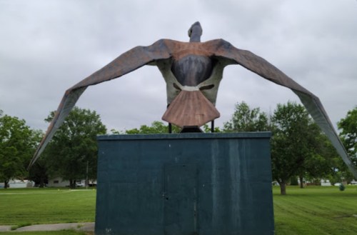 A large metal sculpture of a bird with outstretched wings, perched on a green pedestal in a grassy park.