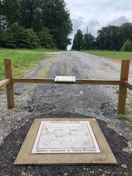 A gravel road leads to a wooden barrier with a plaque marking a surveyor's site in Missouri.