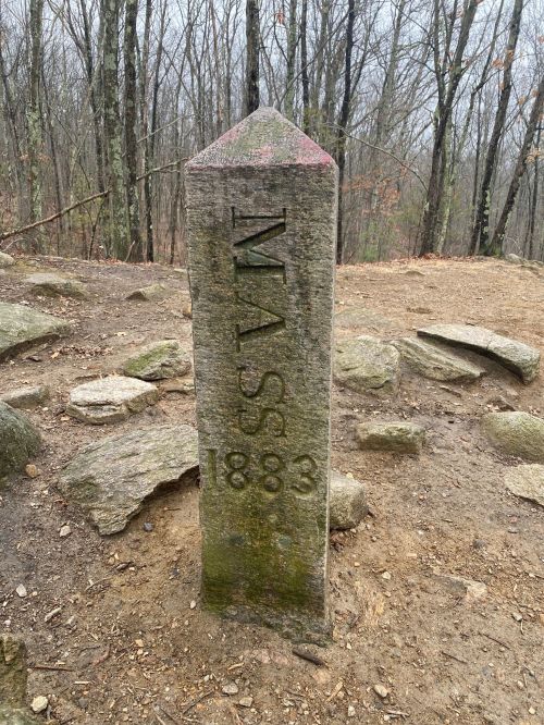 A weathered stone marker inscribed with "MASS" and "1883," surrounded by rocky terrain and bare trees.