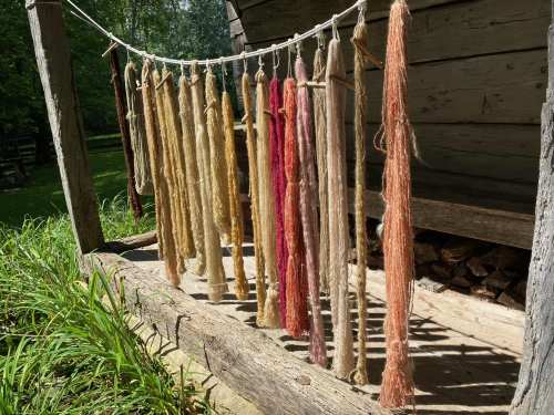Colorful strands of yarn hanging on a line outside a wooden structure, surrounded by greenery.