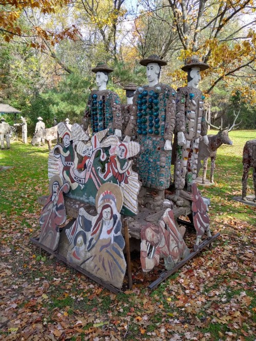 Colorful sculptures of two figures with hats, surrounded by painted scenes, set in a wooded area with autumn leaves.