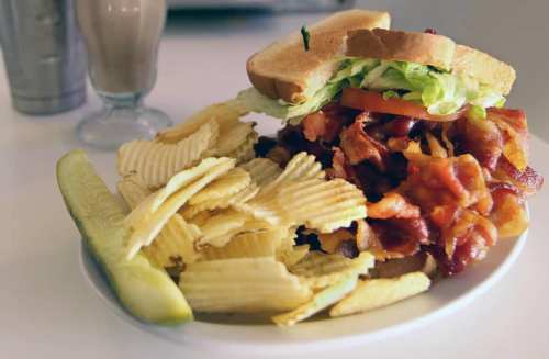 A plate with a towering bacon sandwich, lettuce, tomato, potato chips, and a pickle on the side.