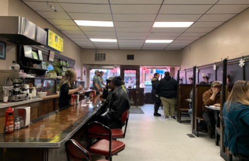 A cozy diner interior with a counter, patrons seated at tables, and a server interacting with customers.
