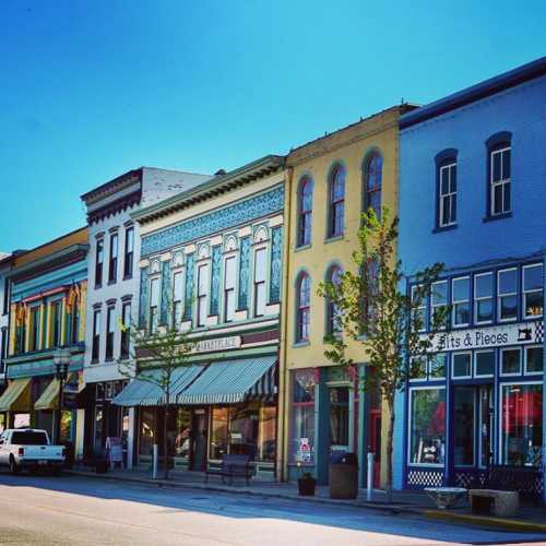 Colorful storefronts line a quaint street, featuring trees and a parked white truck under a clear blue sky.