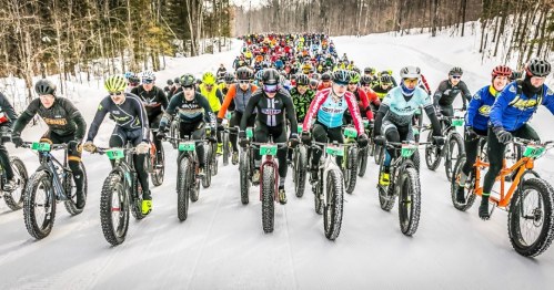 A large group of cyclists in winter gear participating in a snowy fat bike race on a forested trail.