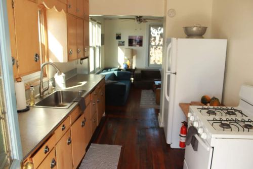 Bright kitchen with wooden cabinets, a sink, stove, and white fridge, leading to a cozy living area with blue couches.