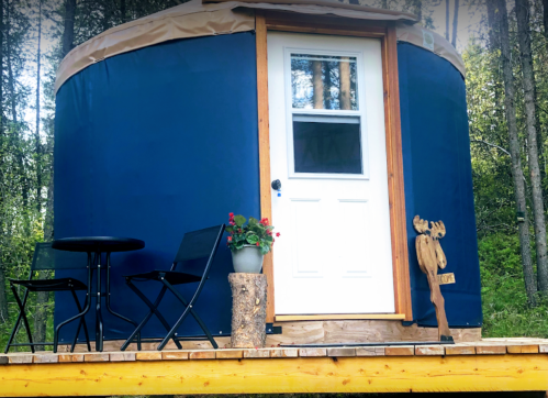 A cozy blue yurt with a white door, surrounded by trees, featuring a small patio with chairs and a potted plant.